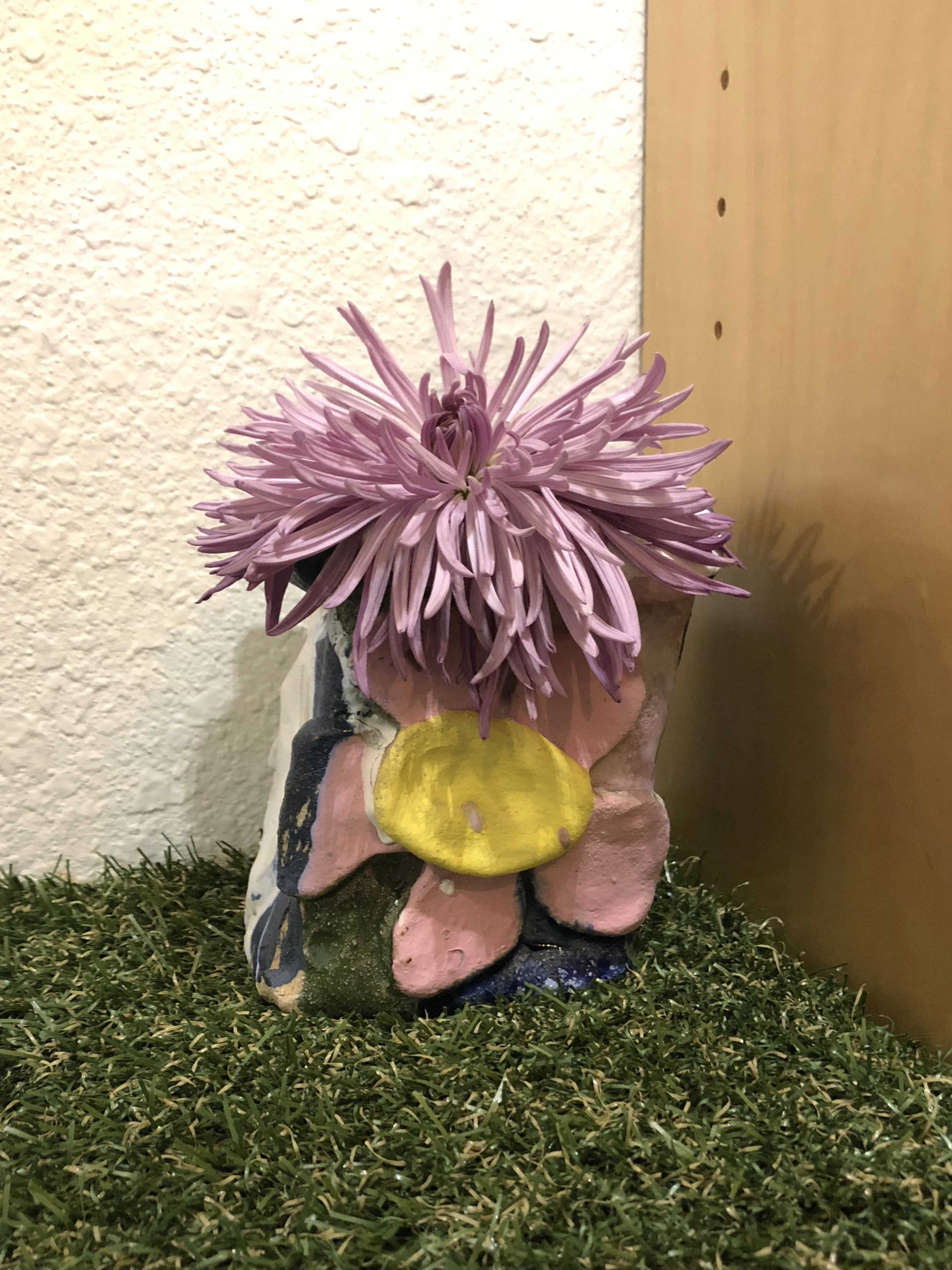 A small, colorful ceramic vase with a pink flower in it on astroturf